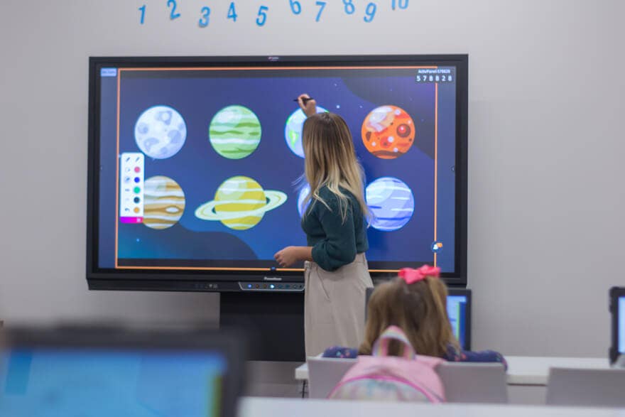 Teacher pointing at an interactive display of planets during a classroom activity, with students working at desks in the foreground.
