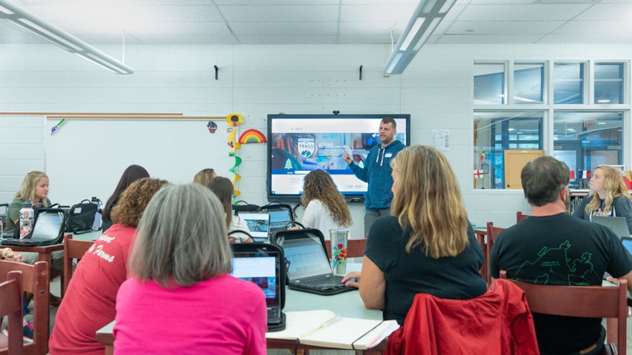 a group of teachers sitting in a classroom setting and listening to a presenter talking to the group